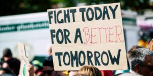 Crowd holding a protest sign with 'Fight Today for a Better Tomorrow', outdoors and during the day.